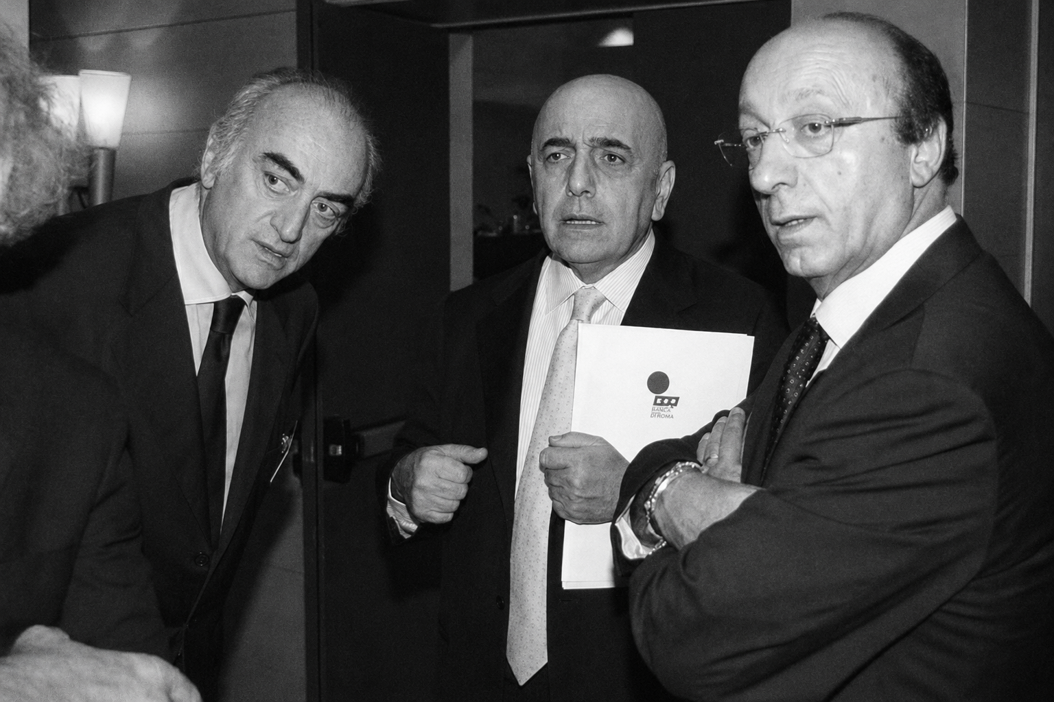 Une photographie argentique en noir et blanc de Luciano Moggi et Adriano Galliani discutant à voix basse dans les tribunes de San Siro, manteaux longs et regards fuyants.